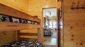 Een houten chalet met stapelbedden, een geruit bankje en een gang die leidt naar een woonruimte met een plafondventilator.
