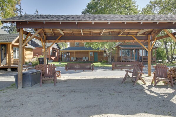 A covered outdoor seating area with wooden chairs and a bench, surrounded by log cabins. - Shawano Lake Cabin w/ Boat Dock! (Cecil)