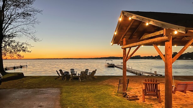 Una terraza de madera con sillas y un área cubierta, con vista a una superficie de agua tranquila al atardecer.