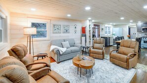 Un salón amplio con un sofá gris, sillones de cuero marrón y un comedor, con una cocina al fondo.