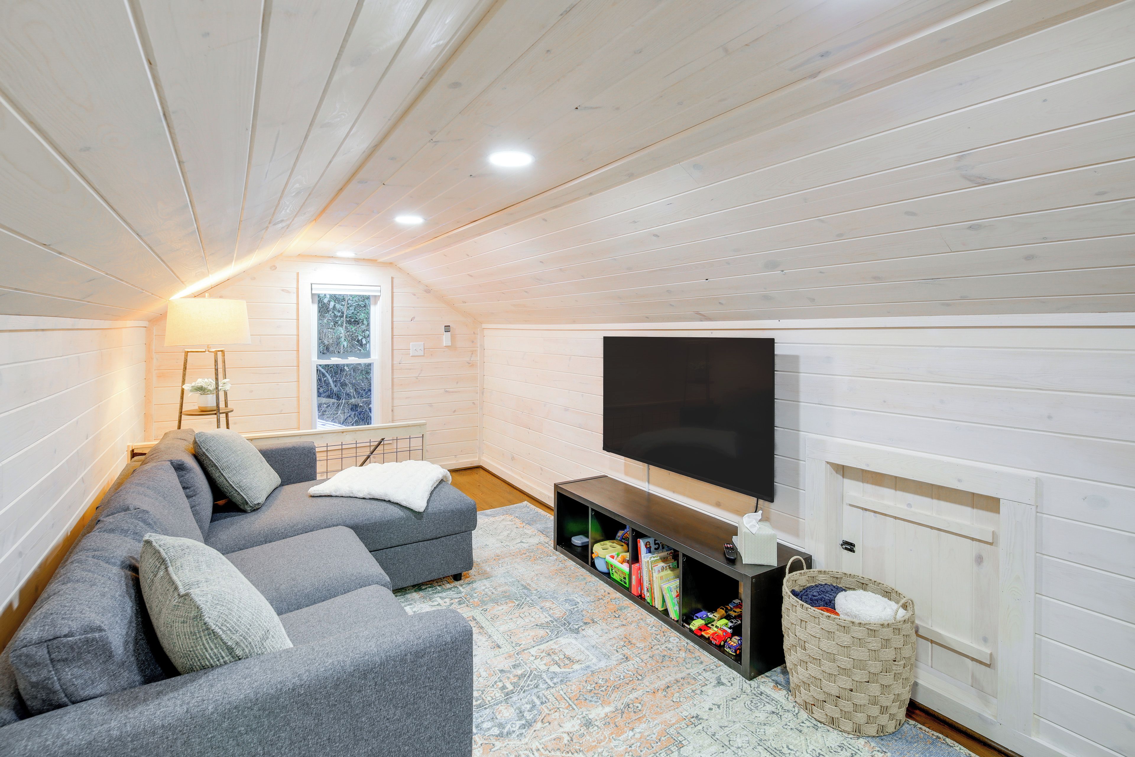 A cozy attic room with a gray sectional sofa, a flat-screen TV, and a small bookshelf.