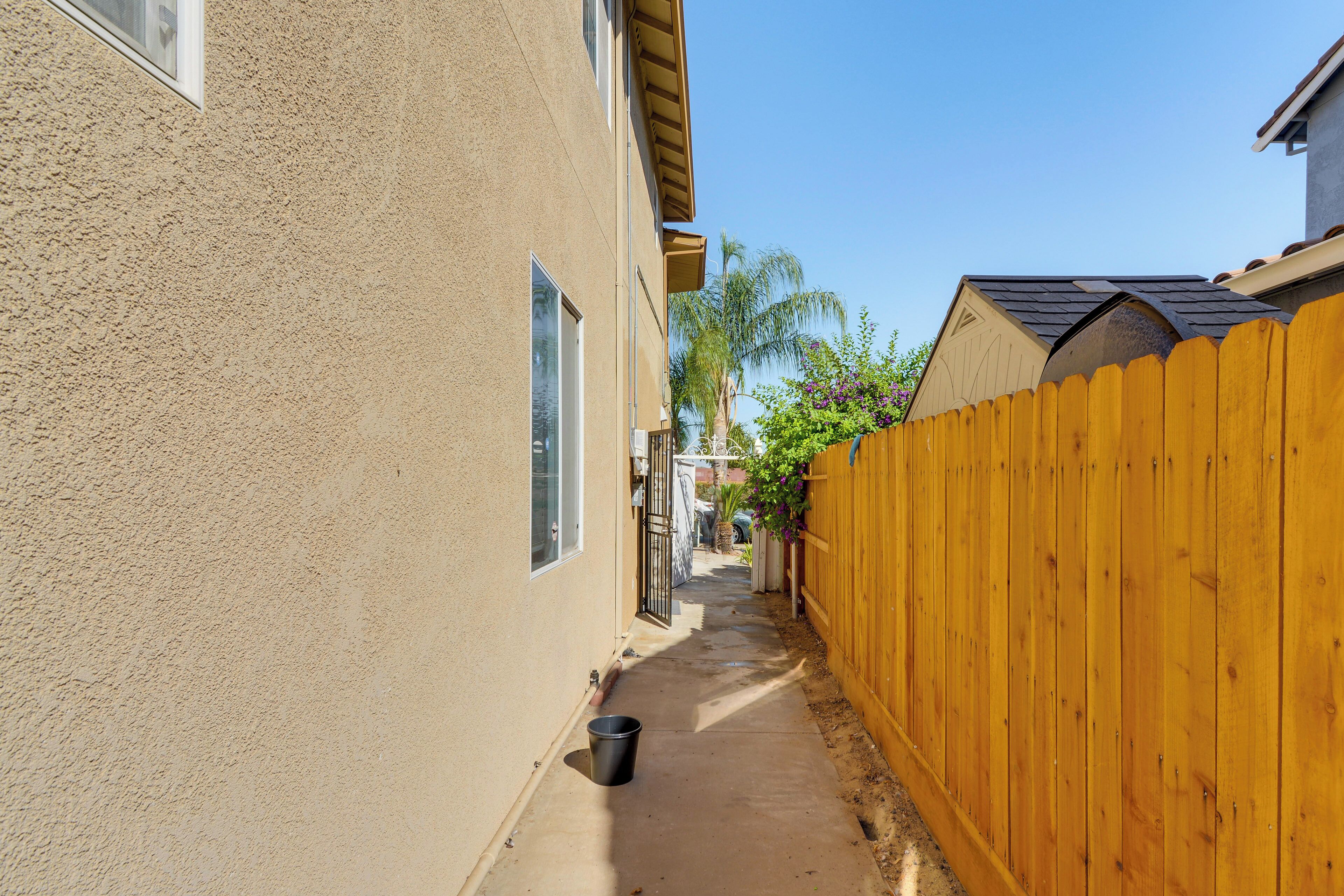 A narrow concrete pathway flanked by a beige stucco building and a wooden fence.