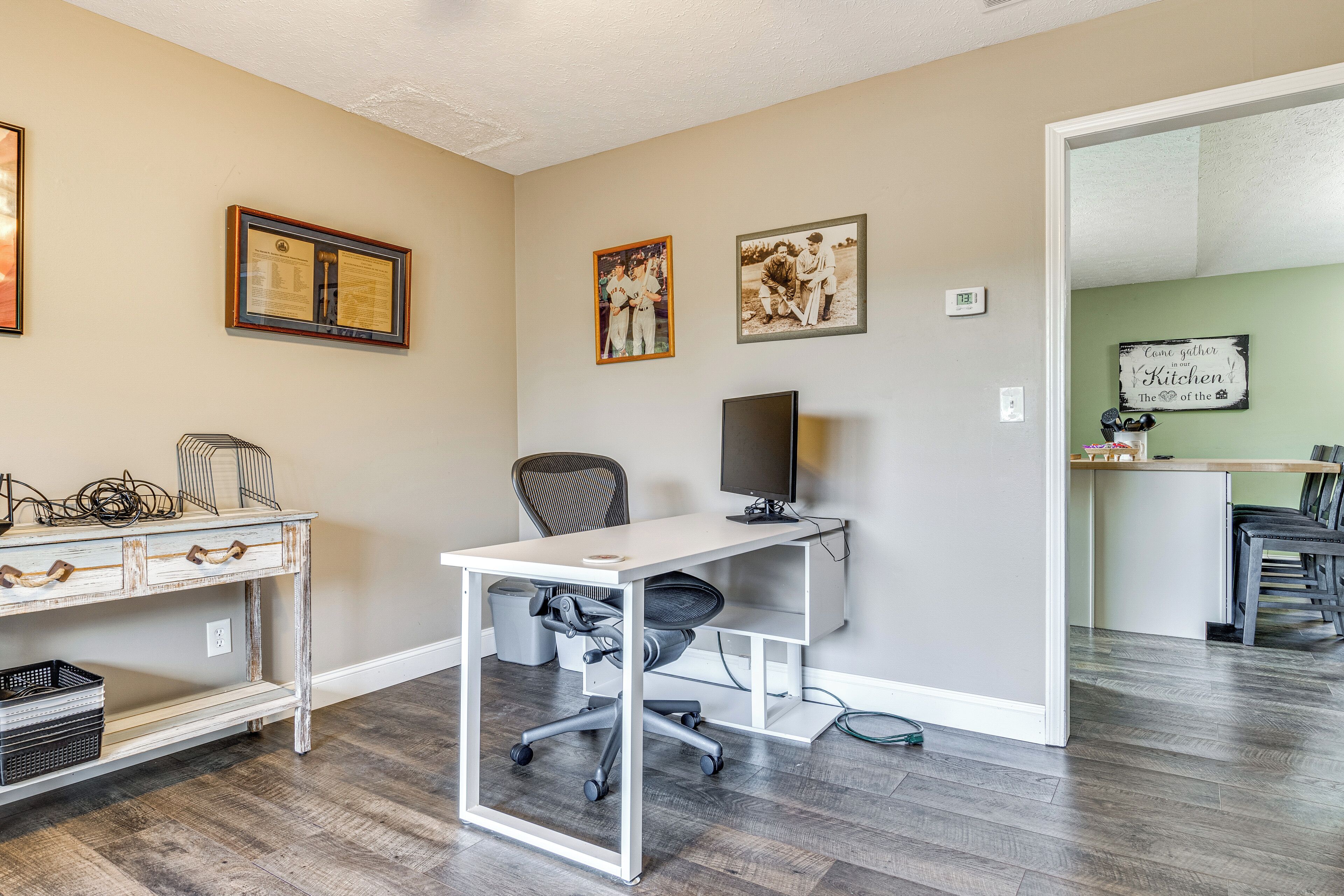 A home office with a desk, chair, and shelves. There are framed pictures on the wall and a sign that says "Come gather 'round the kitchen table."