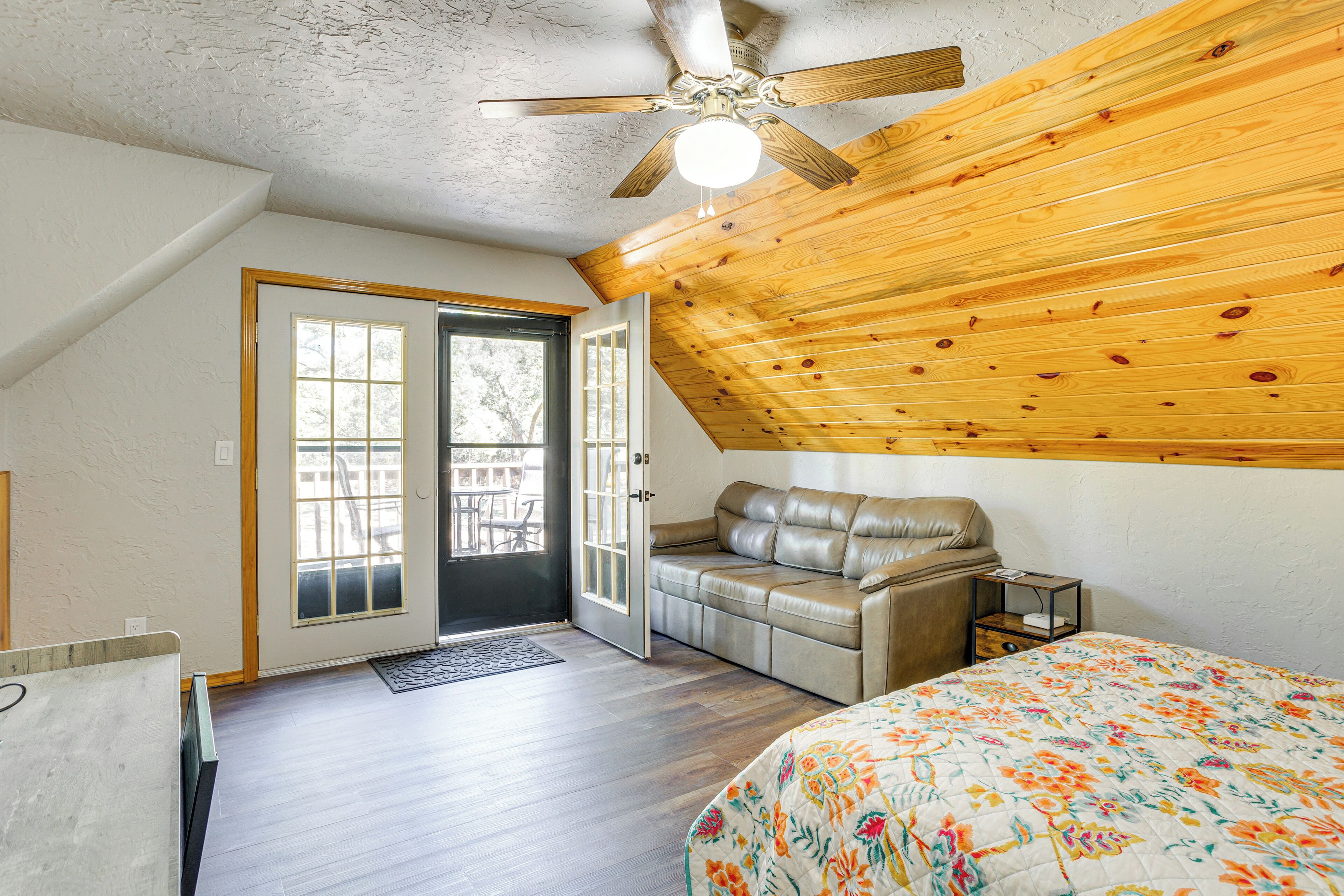 A bedroom with a wooden ceiling, a bed with a floral bedspread, a leather sofa, and a door leading to a balcony.