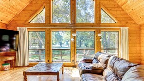 A cozy cabin interior with a wooden ceiling, large windows, a leather sofa, and a wooden coffee table.