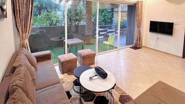 A spacious living area with a large sliding glass door leading to a patio, a sofa, a small table, and a TV mounted on the wall. - Scarlet By Stellar Stays (Mawal)