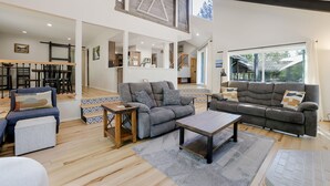A spacious living area with a gray sofa, a blue armchair, and a wooden coffee table. A staircase leads to an upper level with a kitchen and dining area. - NEWB07-Family-Friendly Sunriver Retreat | Hot Tub, A/C, Dog-Friendly, Sleeps 10 (Sunriver)