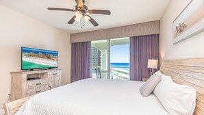 Une chambre d’hôtel avec un grand lit, un ventilateur de plafond, un téléviseur posé sur une commode en bois et une vue sur la plage par la fenêtre.