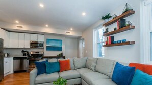A modern kitchen with white cabinets, stainless steel appliances, and a dining area with a grey sofa and colorful cushions. - Boston Apartment | near T (Boston)