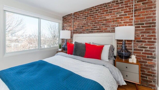 A bedroom with a brick wall, a large window, a bed with a blue blanket, and two bedside tables with lamps. - Boston Apartment | near T (Boston)