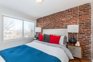 Ein Schlafzimmer mit Backsteinwand, einem großen Fenster, einem Bett mit blauer Decke und zwei Nachttischen mit Lampen.