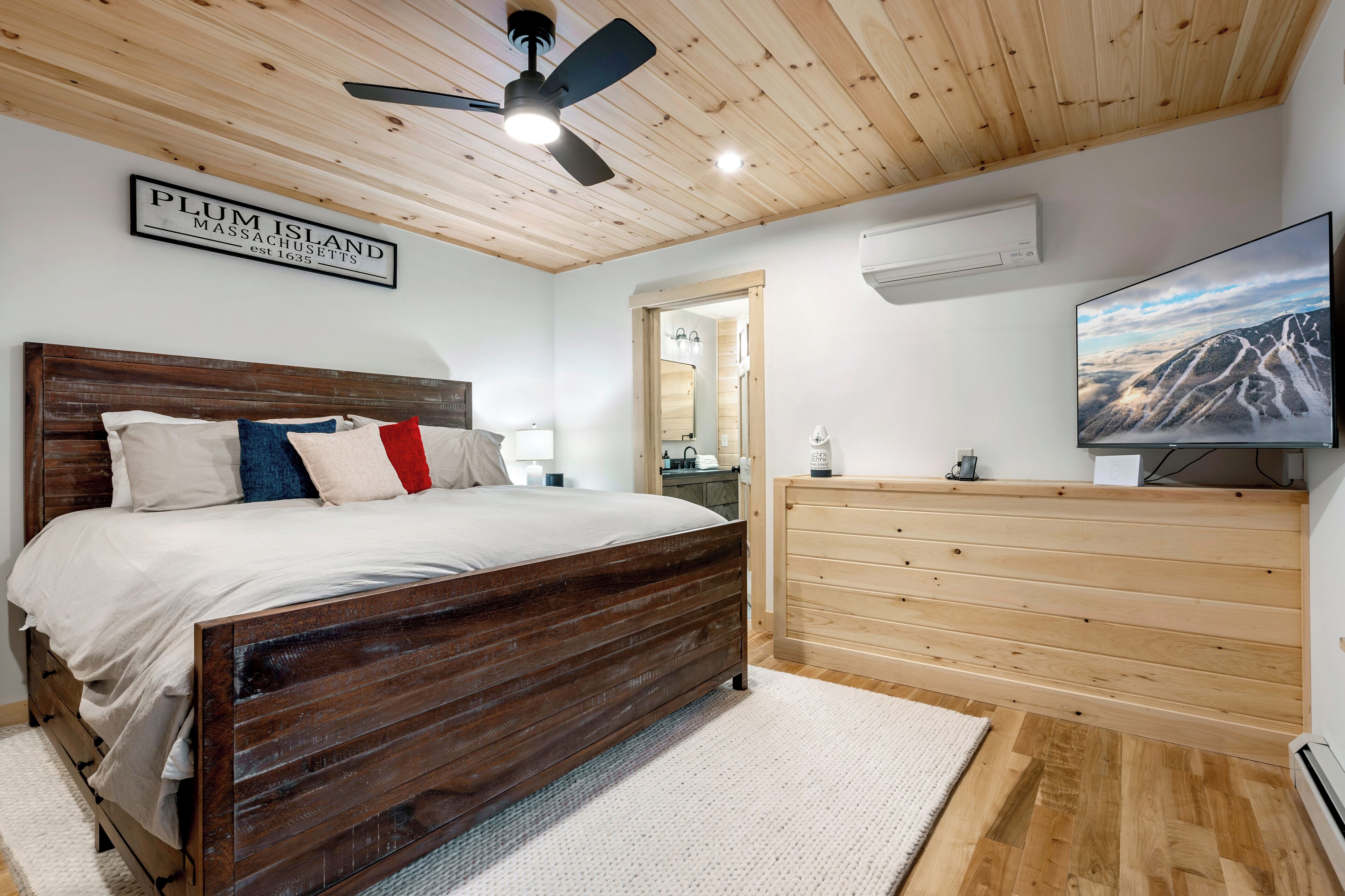 A bedroom with a wooden ceiling, a bed with a headboard, a dresser, a television, and a sign that reads "PLUM ISLAND MASSACHUSETTS."