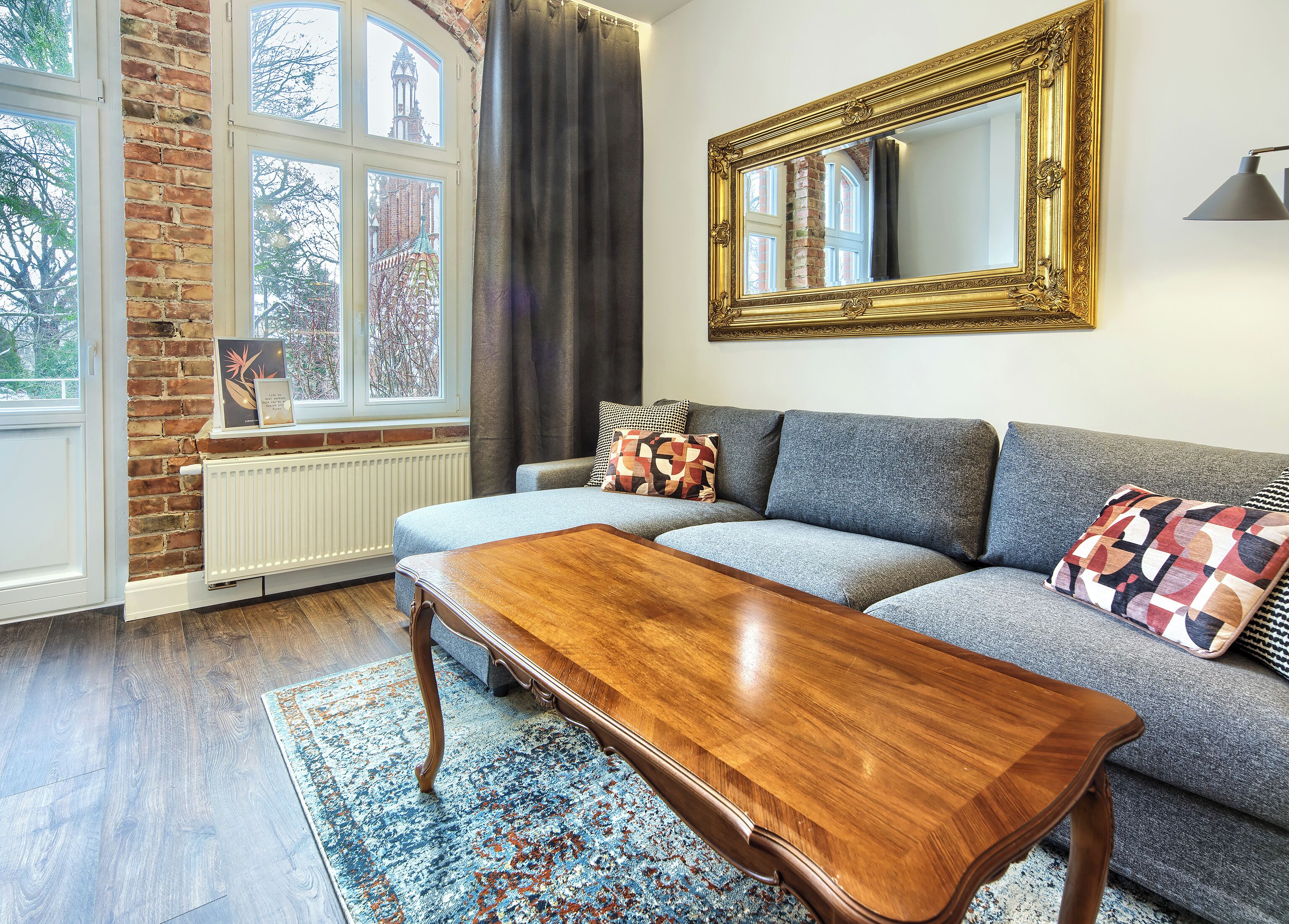 Una sala de estar moderna con una pared de ladrillo, un ventanal grande, un sofá gris, una mesa de centro de madera y una alfombra estampada.