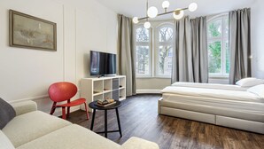A modern hotel room with a large bed, a TV on a white cabinet, a red chair, and a small table with books.