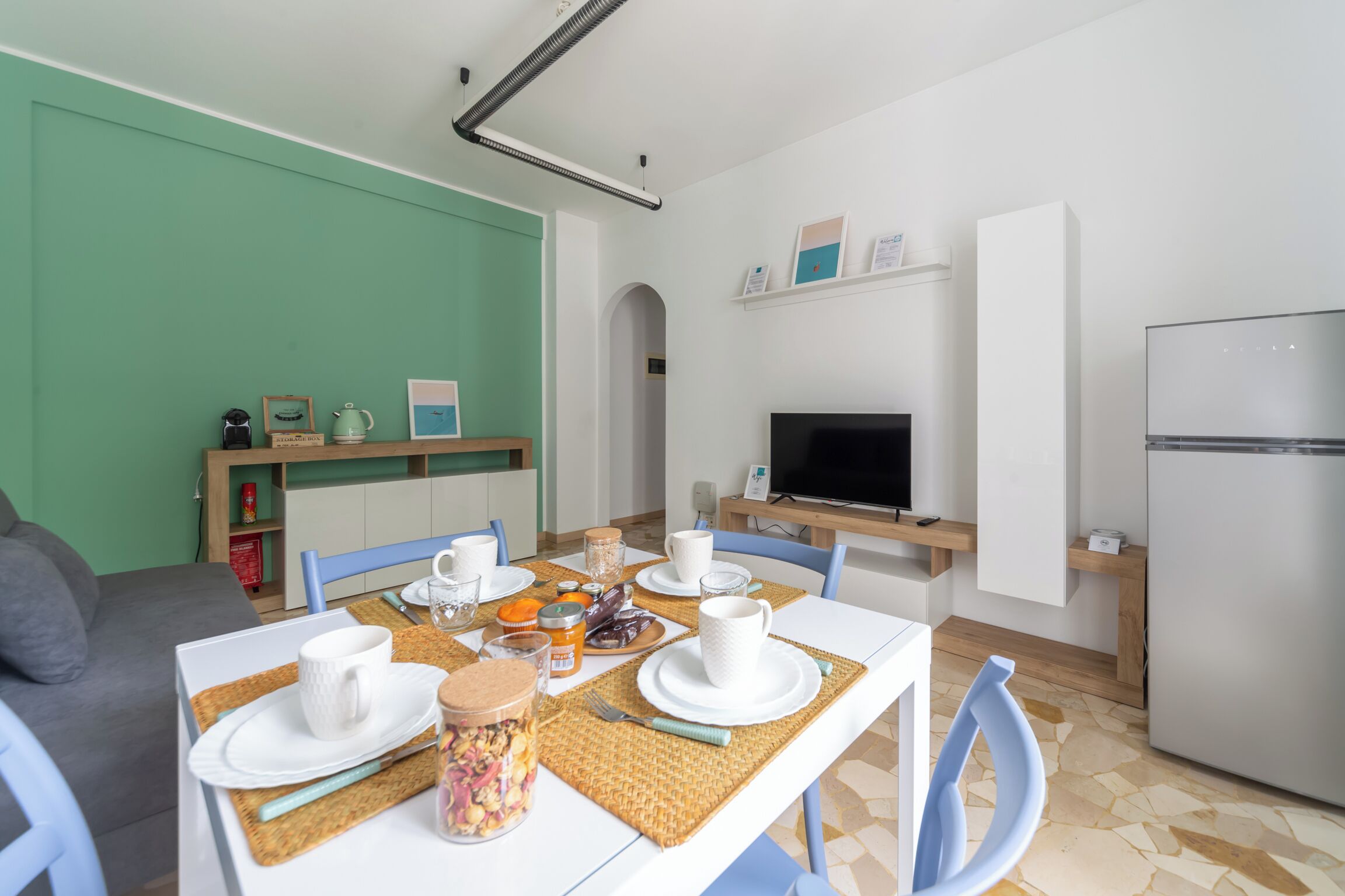 A modern kitchen with white appliances, a dining table set with white plates and cups, and a TV on a shelf.