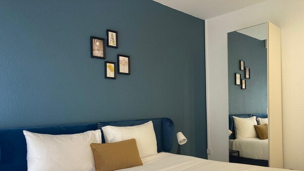 Ein Hotelzimmer mit einem großen Bett, weißen Laken, einem blauen Kopfteil, einem braunen Kissen und einem Spiegel, der den Raum reflektiert.