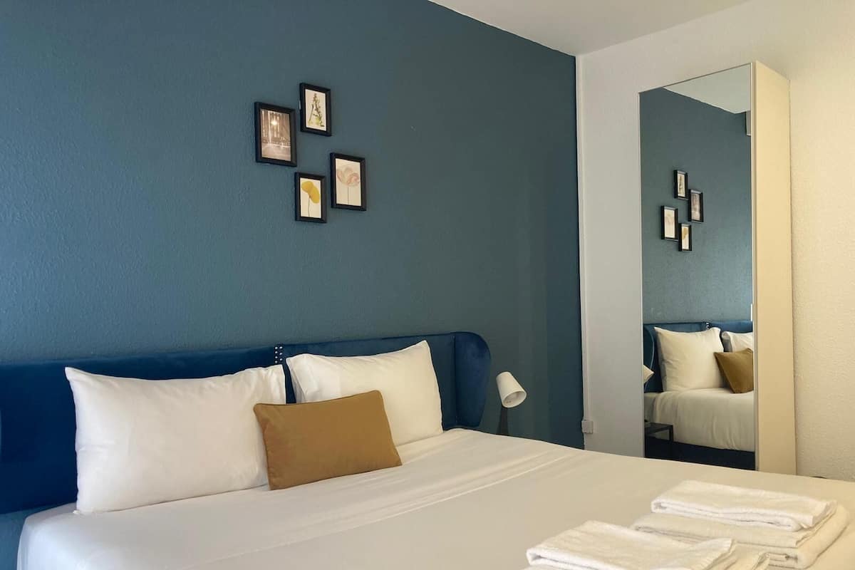 Habitación de hotel con una cama grande, ropa blanca, cabecera azul, almohada marrón y un espejo que refleja la habitación.