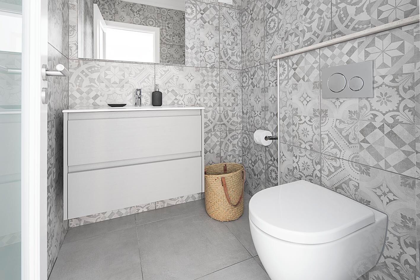 A modern bathroom with patterned tiles, a white toilet, a white cabinet with drawers, and a small trash bin.