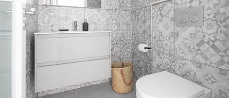 A modern bathroom with patterned tiles, a white toilet, a white cabinet with drawers, and a small trash bin.