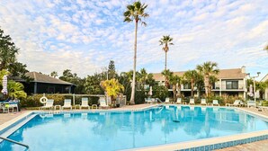 Une piscine rectangulaire dont la profondeur est indiquée par un repère à 1,80 mètre, entourée de chaises longues et de palmiers, avec un bâtiment visible au loin.