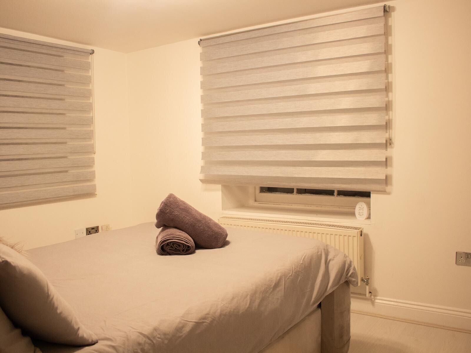 A bedroom with a bed, two windows with blinds, a radiator, and a rolled-up towel on the bed.