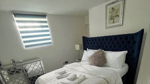 Una cama bien hecha con sábanas y toallas blancas, un cabecero tapizado en azul oscuro, una mesita de noche con lámpara y una ventana con persianas.