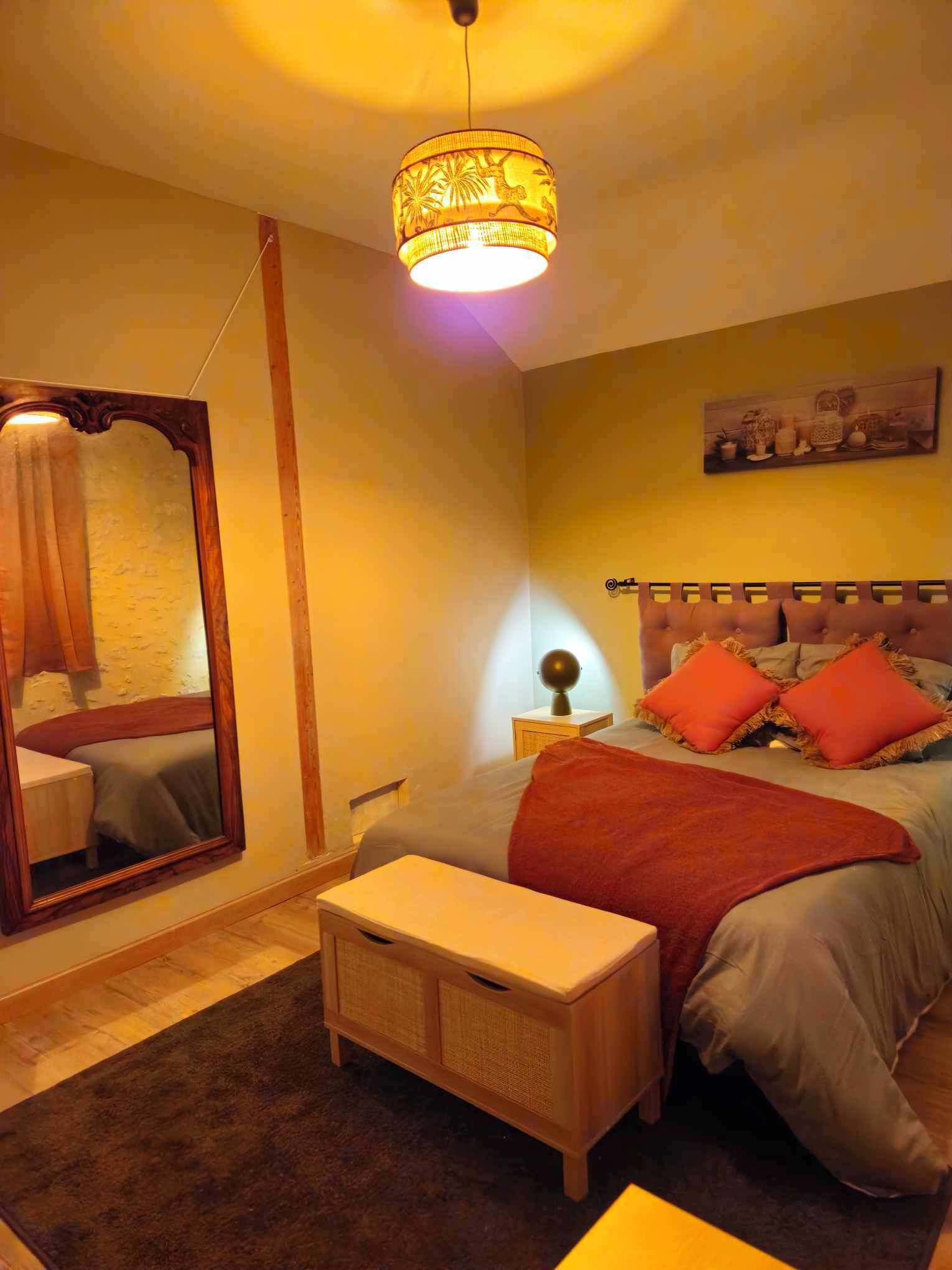 Quarto com cama, mesa de cabeceira, luminária, espelho e luminária pendente.