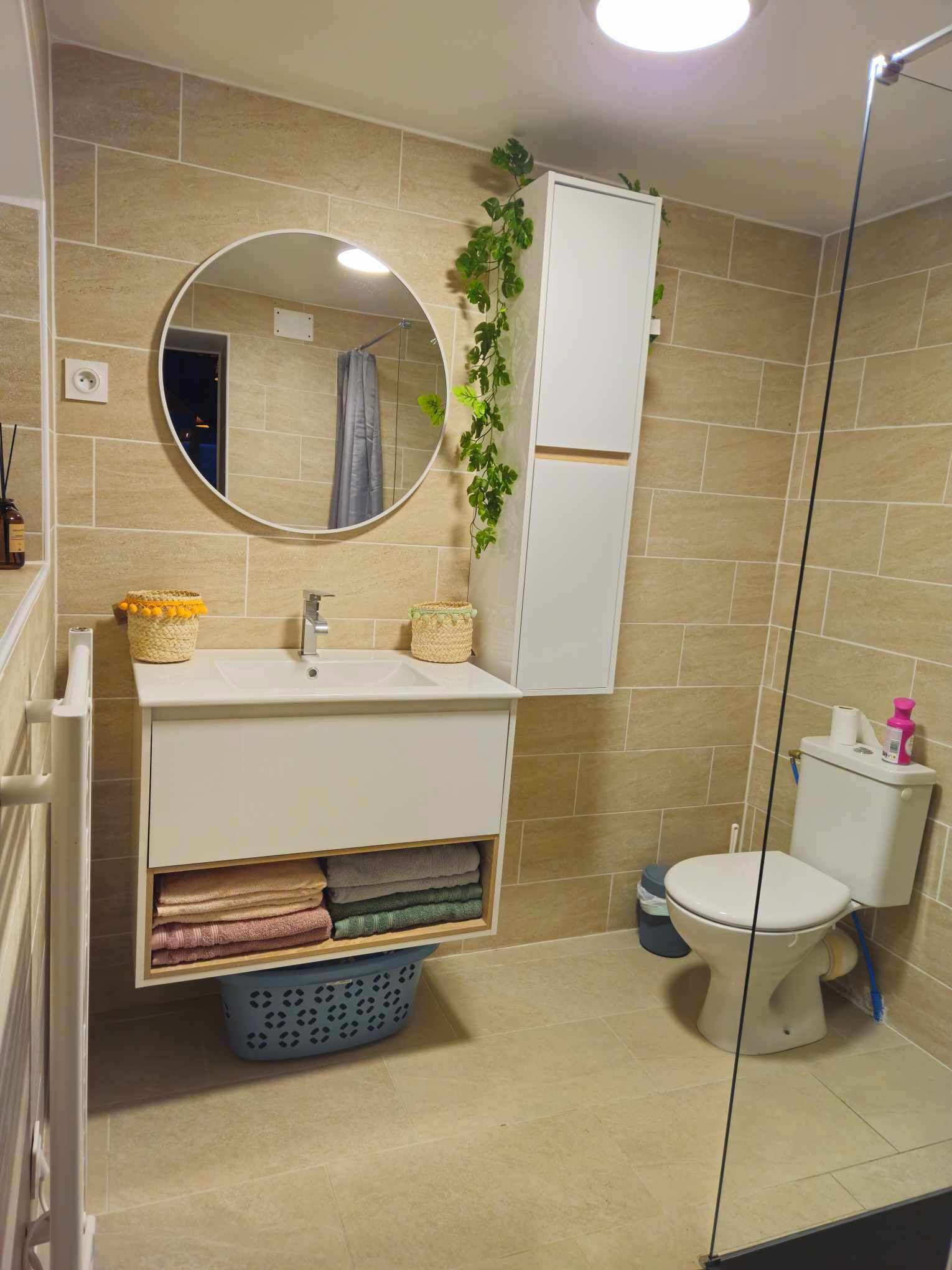 Een badkamer met een witte wastafel, een ronde spiegel, een toilet en een blauwe wasmand.
