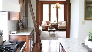Una cocina con estufa a gas, armarios de madera y encimera de mármol. Una escalera conduce a una sala de estar con un sofá, una mesa de centro y una araña.