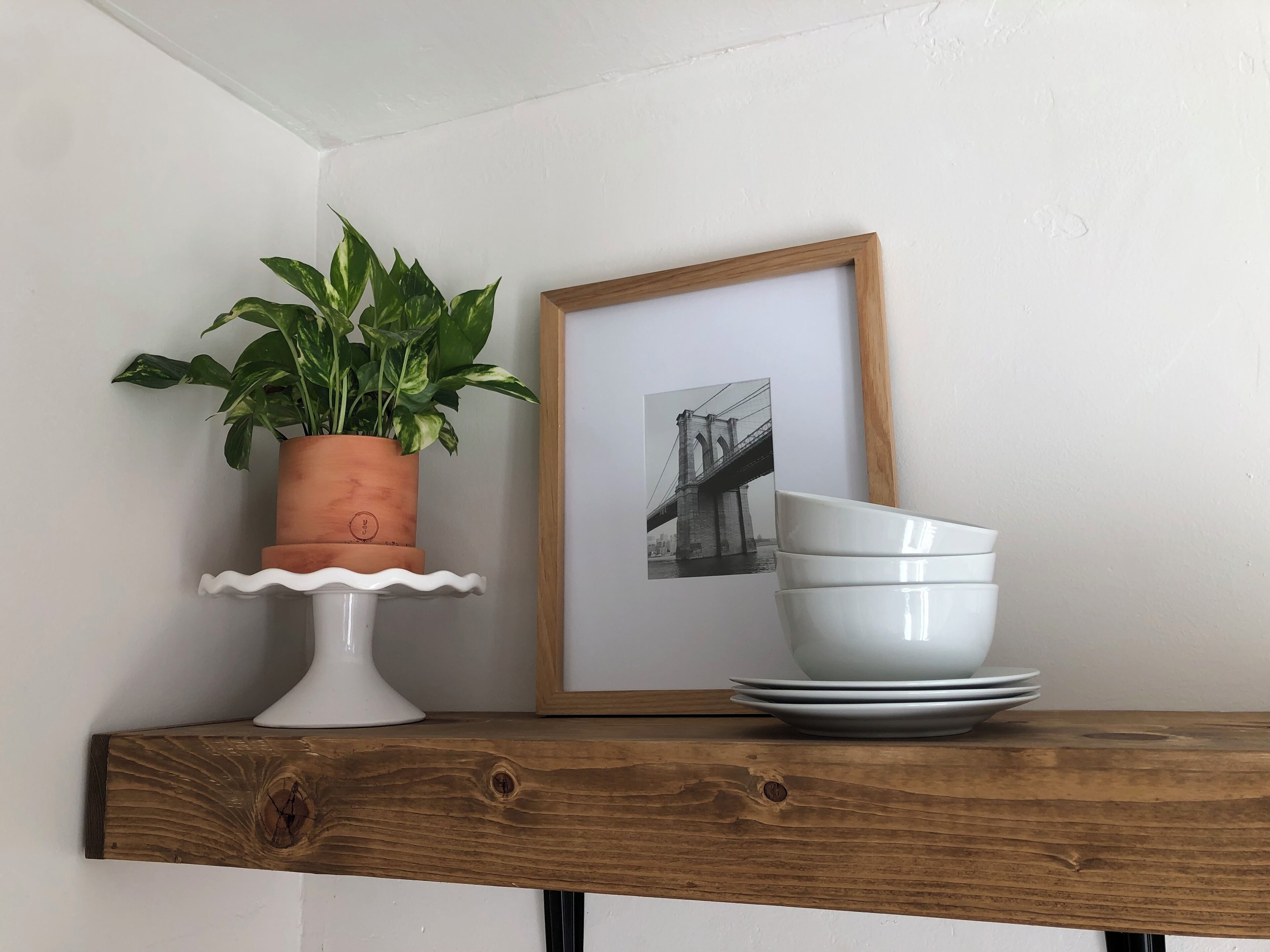 Une étagère en bois avec un pot de plante, un tableau encadré et des bols blancs empilés.