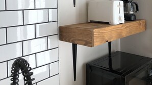 Una cocina con salpicadero de azulejos blancos, un estante de madera, un tostador blanco, una cafetera negra y un microondas negro.