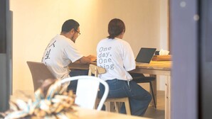 Two people working at a wooden desk with a laptop, in a room with a plain wall and a door in the background. - One Miyakojima (Miyakojima)