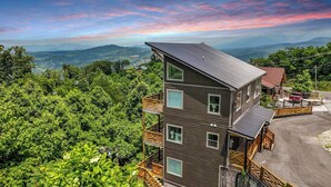 A multi-story building with a balcony, surrounded by lush greenery and mountains in the background.