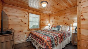 A wooden cabin bedroom with a bed, a nightstand, a lamp, and a window with blinds. - Charming Dog Friendly Retreat w/ Hot Tub (McHenry)