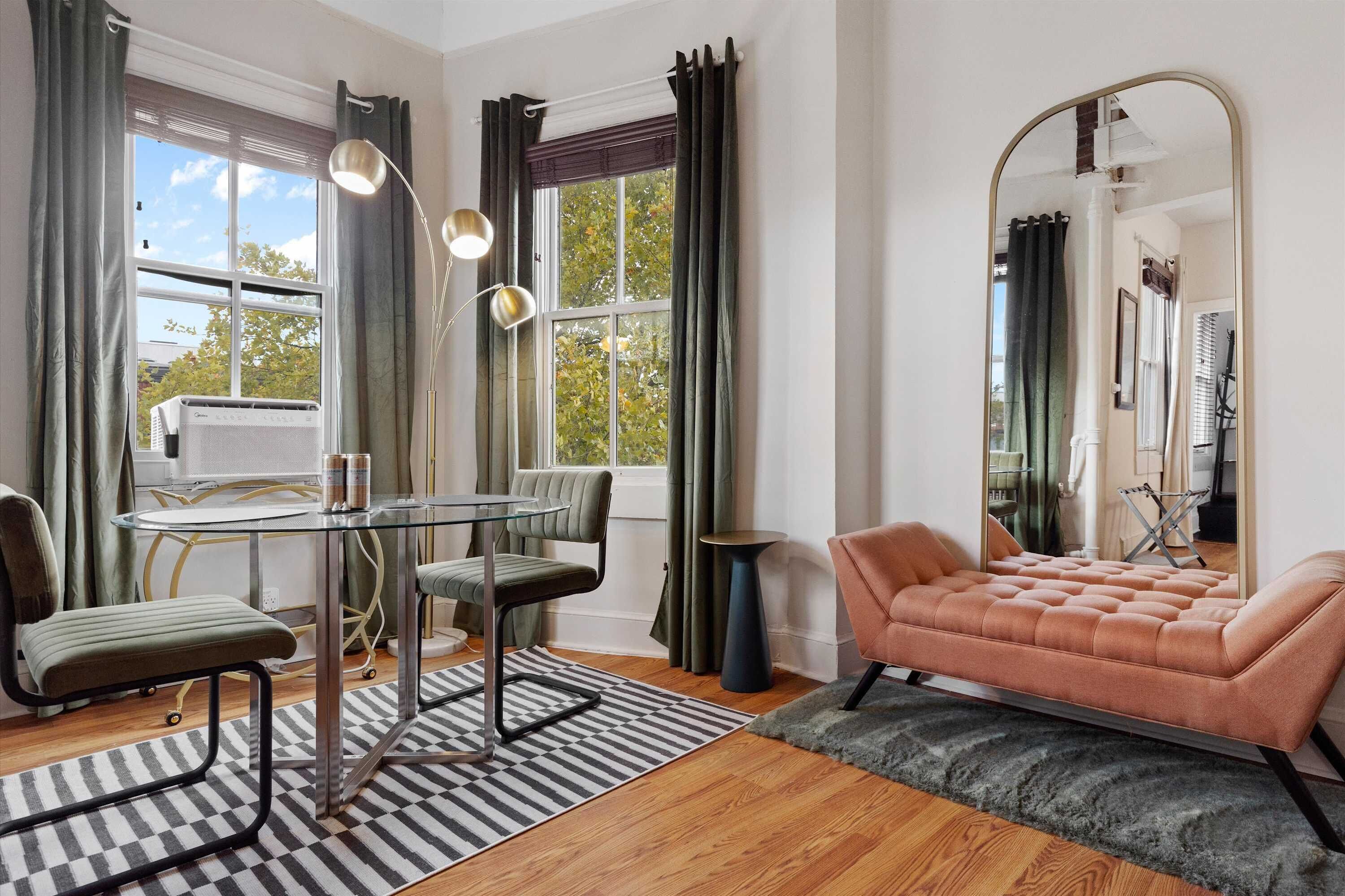 A room with a glass table, chairs, a sofa, a large mirror, and a window with curtains.