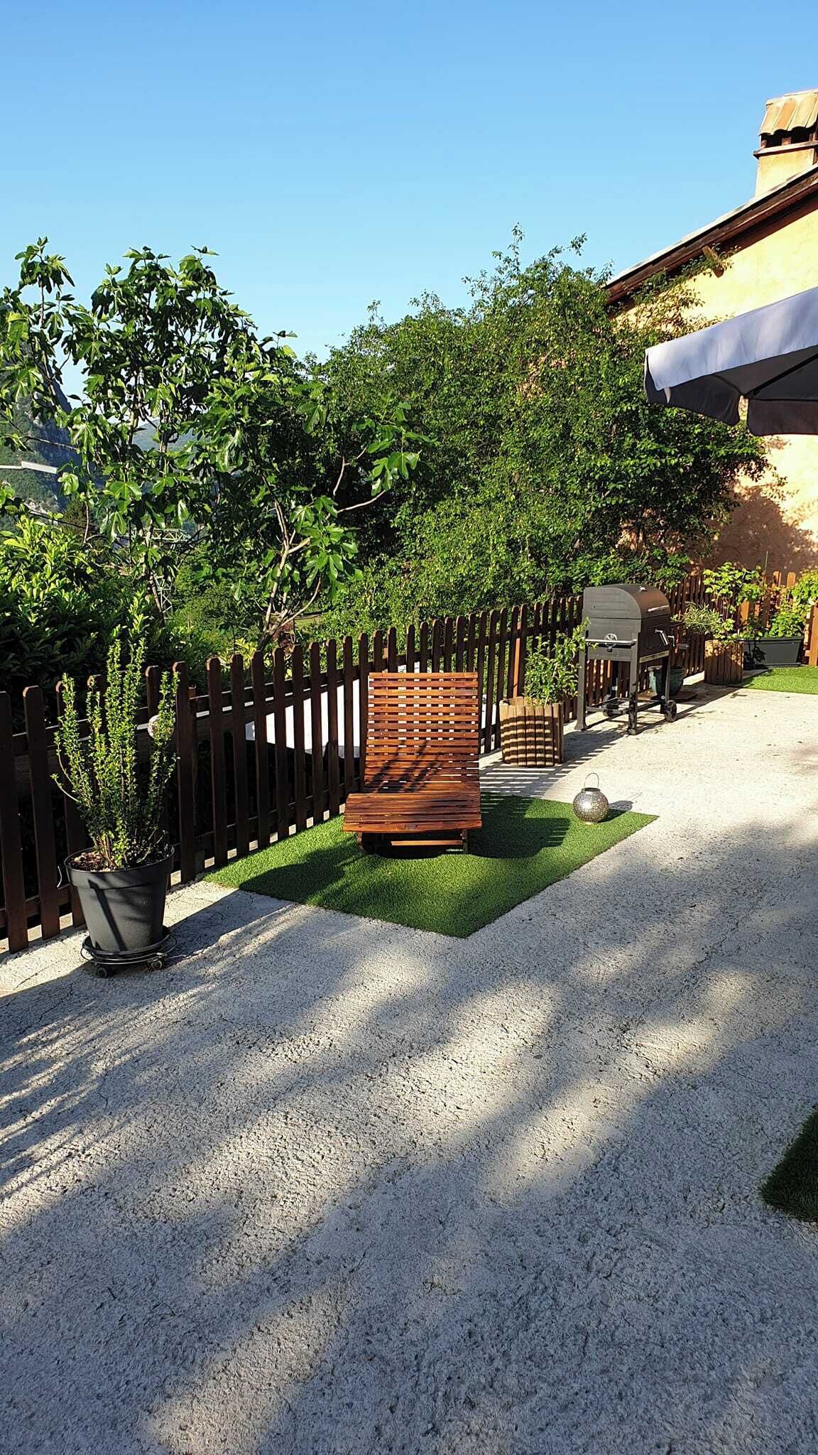 Un chemin pavé mène à un banc en bois, à proximité duquel se trouve un chariot-barbecue. L’ensemble est entouré d’une clôture en bois et d’une végétation luxuriante.