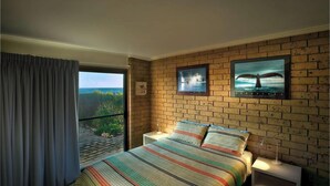 A bedroom with a brick wall, a bed with a colorful striped bedspread, two framed pictures of whales, a window with a view of the sea, and a curtain partially drawn. - Absolute Beachfront Port Elliot | Ocean Views | Sleeps 8 | Family Holiday Accommodation (Port Elliot)