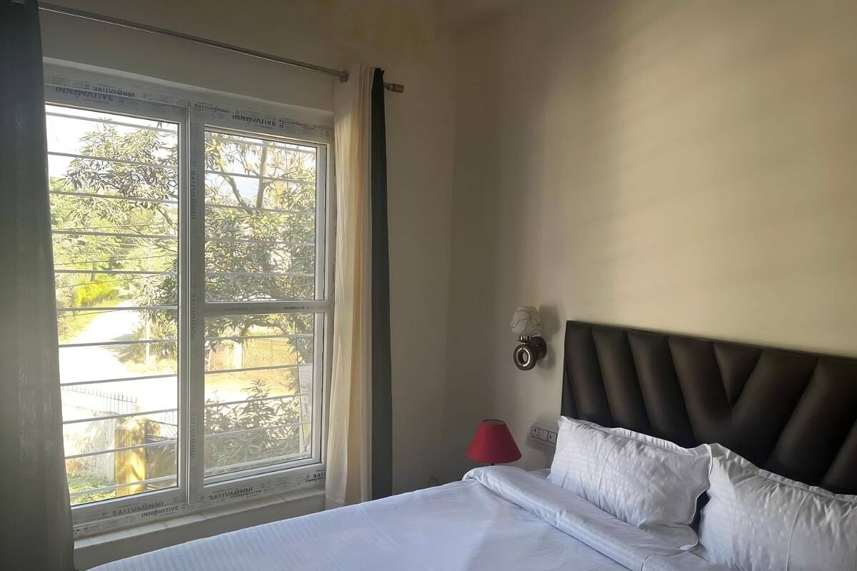 A hotel room with a bed, a window with curtains, a wall-mounted lamp, and a view of greenery outside.