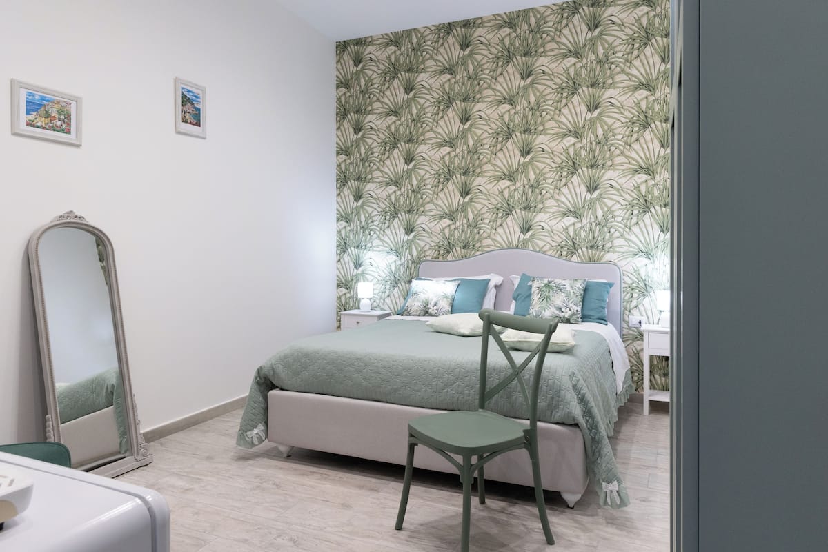 A bedroom with a bed, a chair, a mirror, and a wall with a leafy pattern.