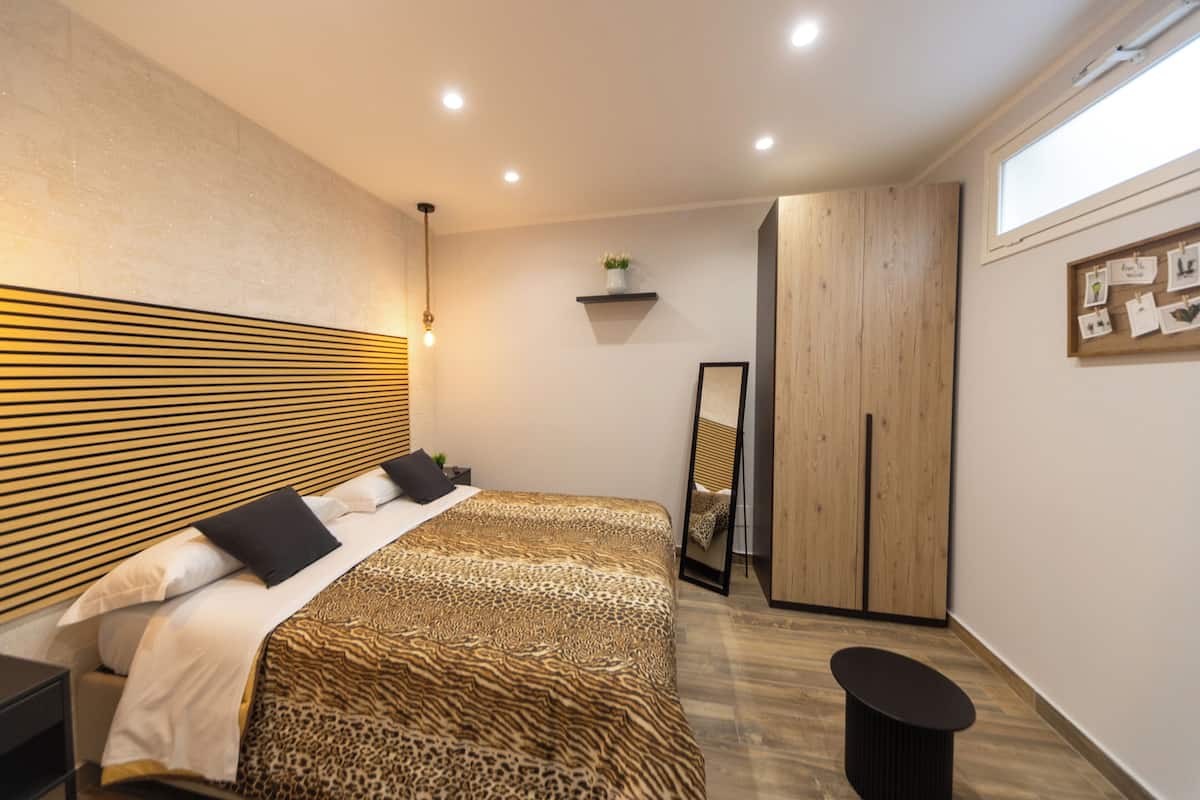 A modern bedroom with a wooden headboard, a bed with a leopard print bedspread, a bedside table, a mirror, a wardrobe, and a small shelf with a plant.