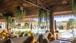 Un restaurant doté d’une terrasse avec vue sur une piscine, agrémentée de plantes suspendues et surmontée d’un toit de chaume.