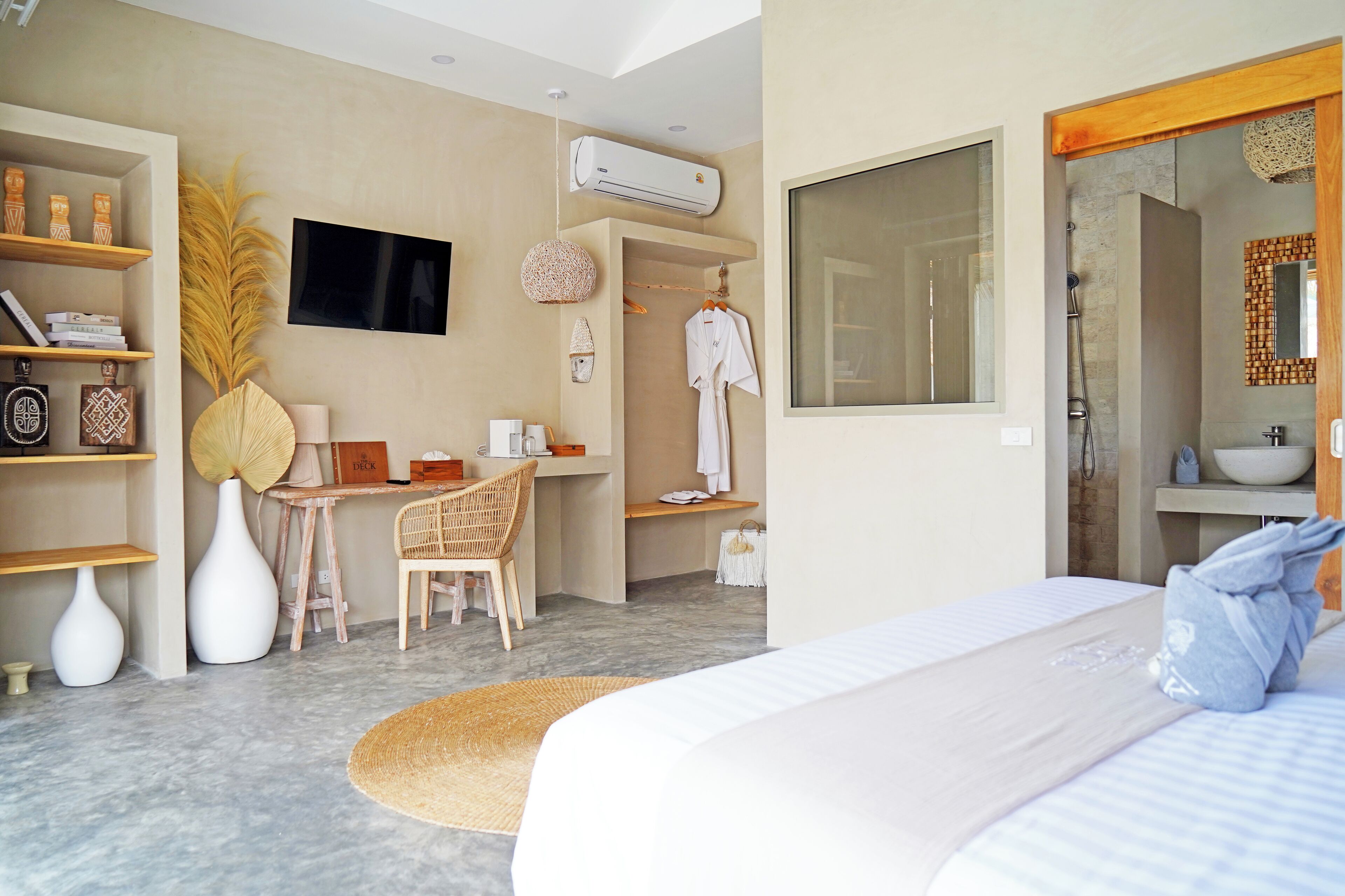 A modern hotel room with a bed, a desk, a chair, a TV, an air conditioning unit, and a bathroom with a shower and sink.