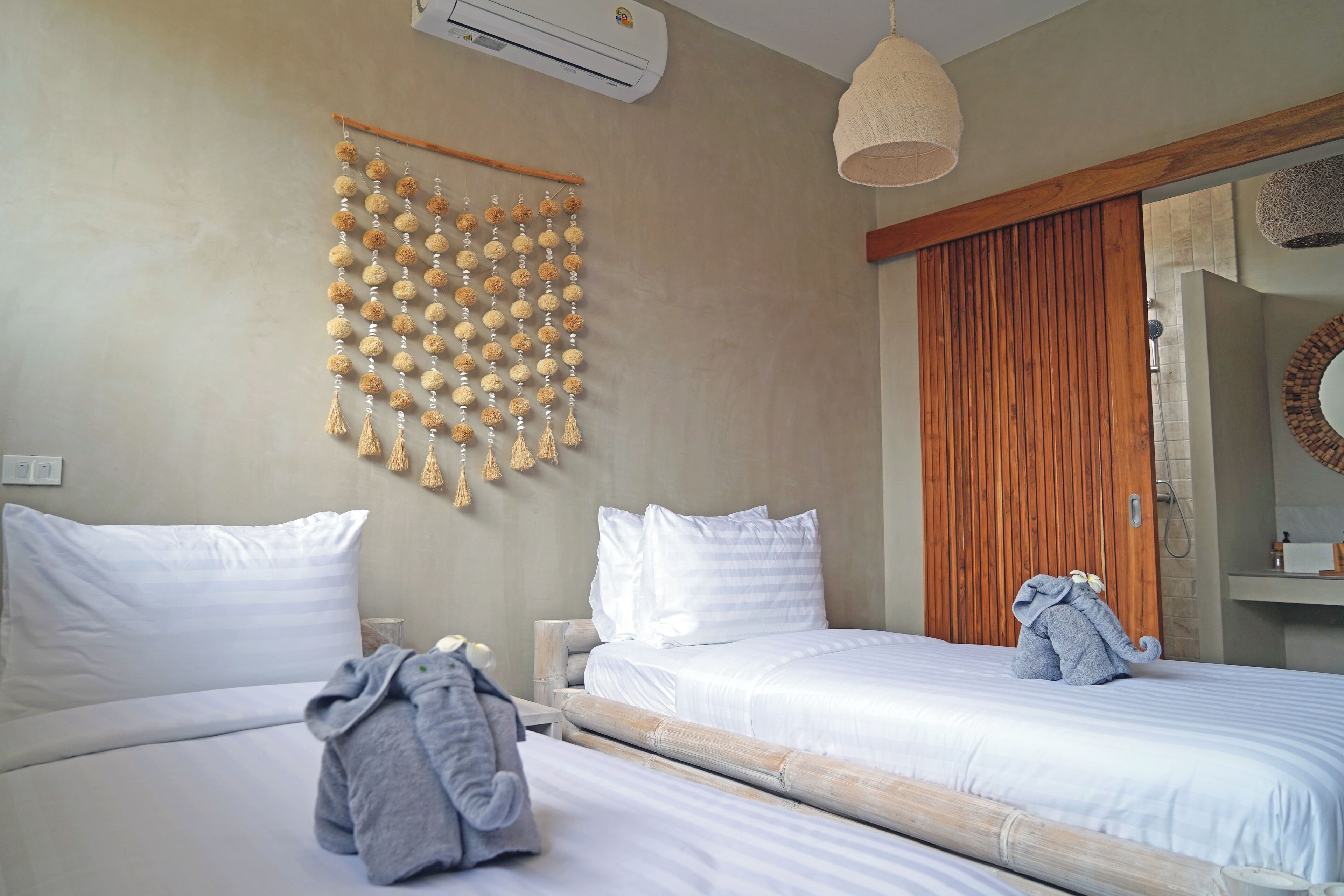 A hotel room with two beds, a wall-mounted air conditioner, a wooden slat door, and a hanging decorative piece.