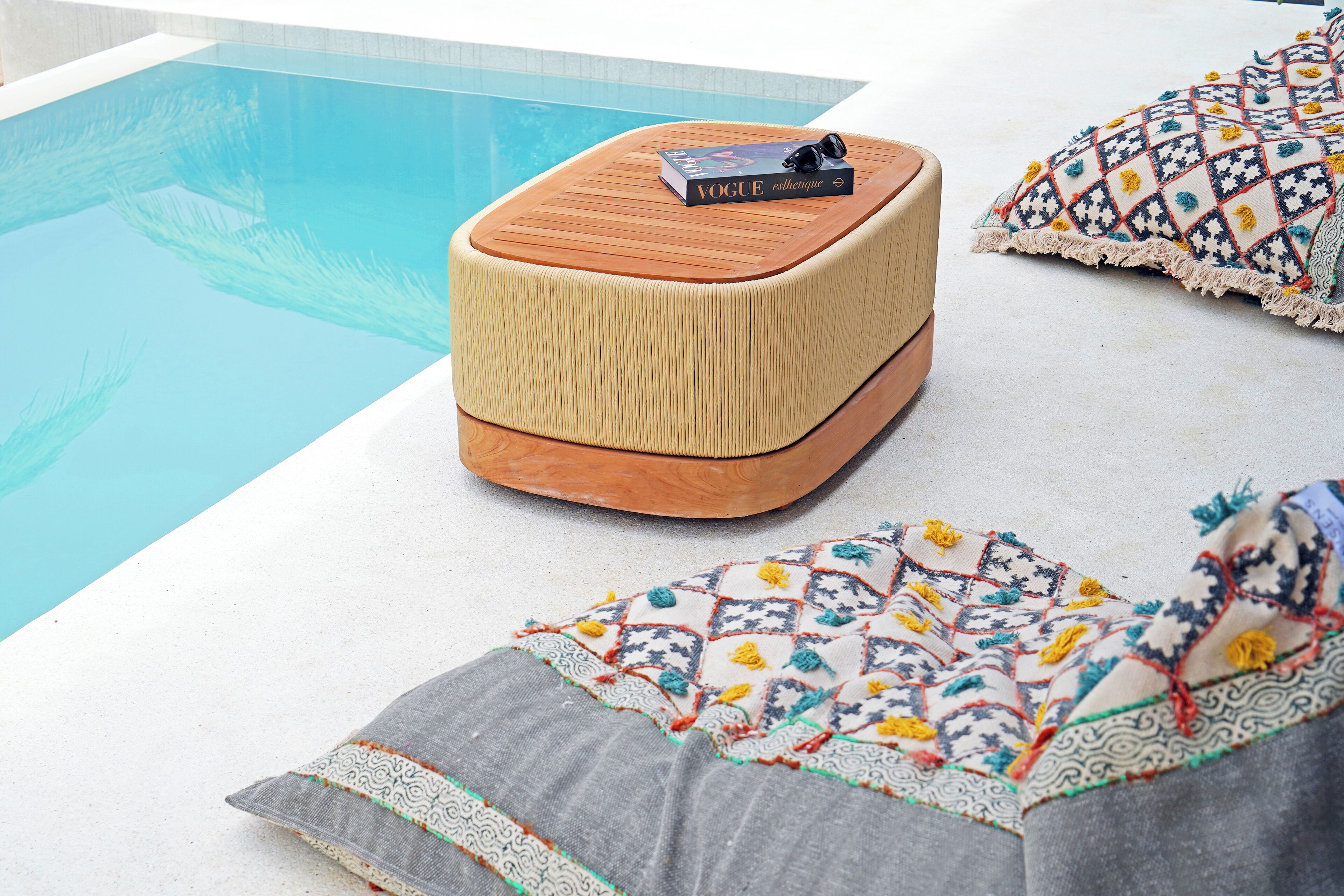 Un área junto a la piscina con un taburete de madera, un libro y un cojín estampado.