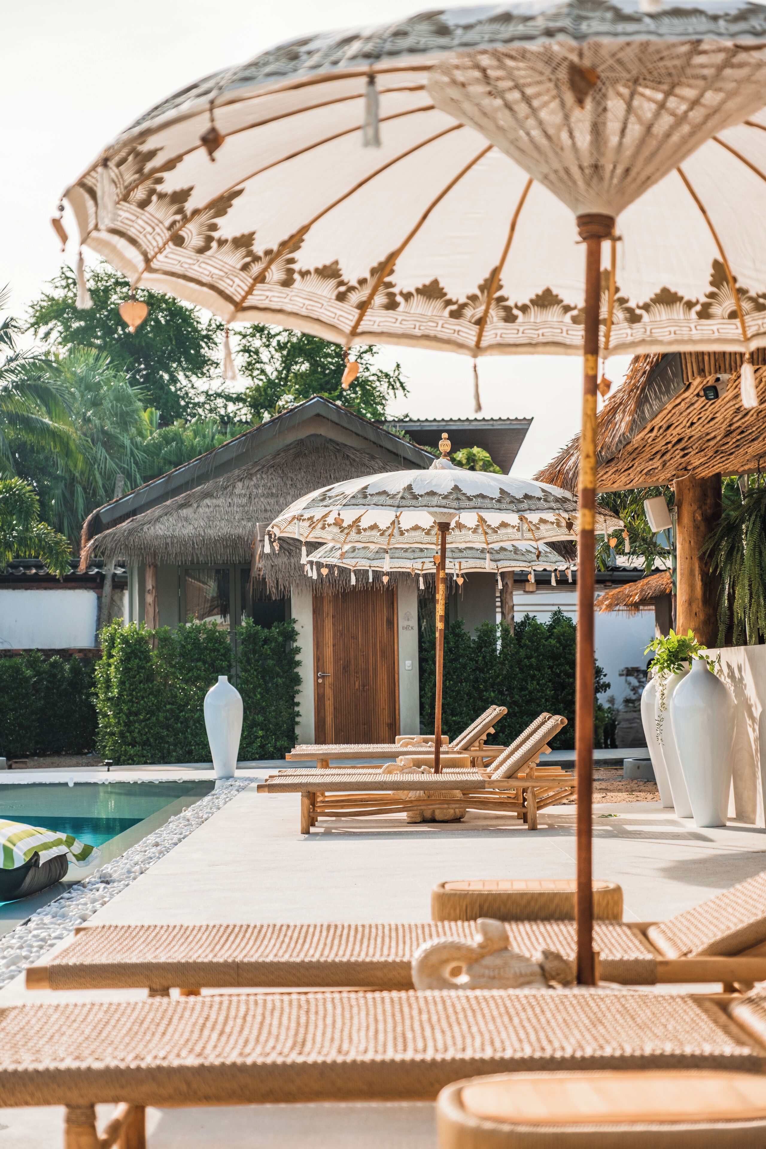 A poolside area with lounge chairs, a decorative umbrella, and a thatched-roof building.