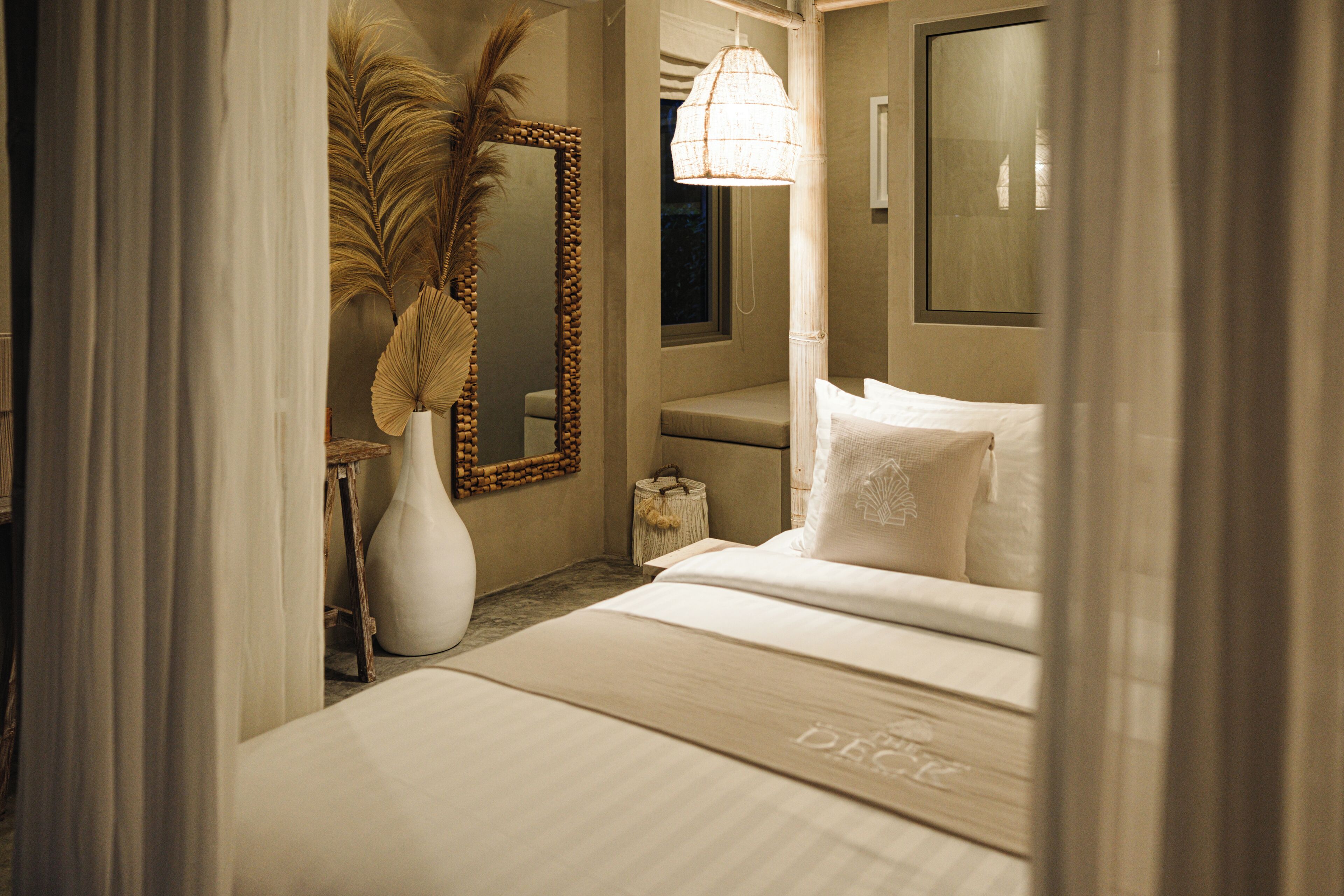 A hotel room with a bed, a mirror, a vase with dried plants, and a hanging light fixture.