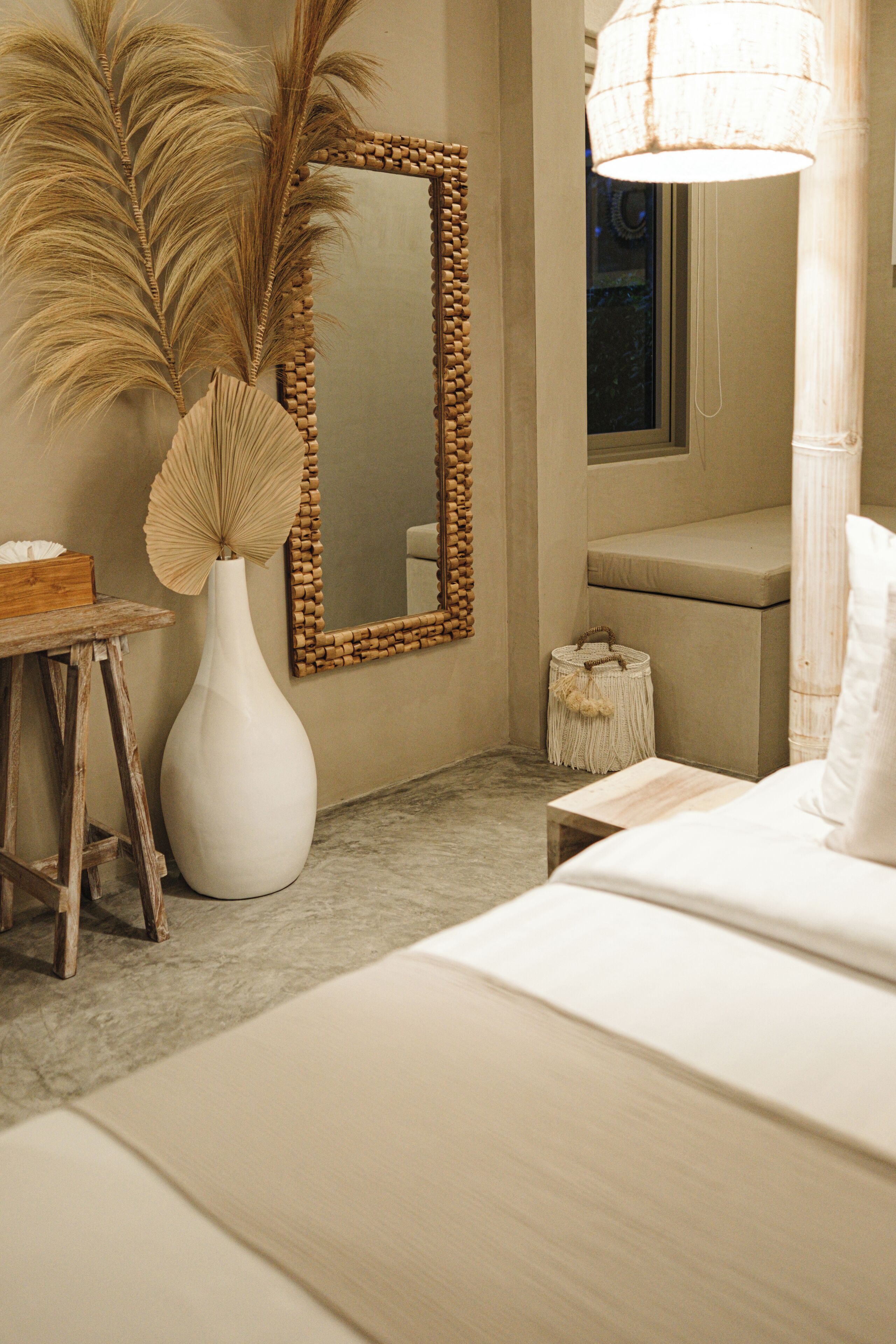 A bedroom with a bed, a large mirror, a vase with dried plants, and a bedside table.