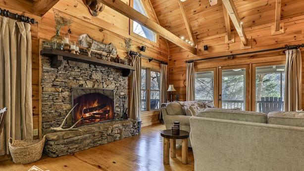 A cozy cabin interior with a stone fireplace, wooden beams, a sofa, and large windows. - Sweet Water Retreat | No Pet Fees | Sleeps 5 | Coosawattee Riverfront (Ellijay)
