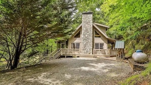 A cabin with a stone chimney, wooden deck, and a gravel driveway surrounded by trees. - Sweet Water Retreat | No Pet Fees | Sleeps 5 | Coosawattee Riverfront (Ellijay)