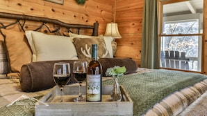 A wooden cabin room with a bed, a tray with wine glasses and a bottle, and a window with a view of trees. - Sweet Water Retreat | No Pet Fees | Sleeps 5 | Coosawattee Riverfront (Ellijay)